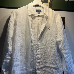 Vit linneskjorta från Polo Ralph Lauren - Klassisk vit skjorta i linne från Polo Ralph Lauren med ärm och knappar framtill. Broderad logga på bröstet. Perfekt för en stilren och avslappnad look.
