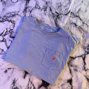 Ljusblå t-shirt från Polo Ralph Lauren - Säljer en ljusblå t-shirt från Polo Ralph Lauren med klassisk passform och kort ärm. T-shirten har en bröstficka med den ikoniska orange broderade loggan. Perfekt basic-plagg med rund hals och mjukt bomullsmaterial. Skriv gärna om man har frågor eller vill förhandla. 