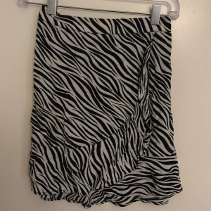 Zebramönstrad kortkjol i svartvitt - Snygg kortkjol med zebramönster i svart och vitt. Kjolen har omlott-look och volangkant nertill som ger en cool och trendig vibe. Perfekt för dig som vill sticka ut med ett djurmönster. Resår i midjan för skön passform.