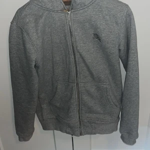 Grå hoodie från Burberry med dragkedja - Grå hoodie från Burberry med klassiskt rutigt foder på insidan och broderad ryttare på bröstet. Tröjan har huva, dragkedja framtill och två fickor. Perfekt för dig som vill ha en stilren och lyxig vardagslook.