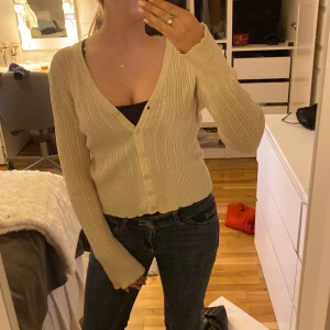 Beige ribbad kofta - Superfin beige kofta med ribbat och kabelstickat mönster. Croppad modell med v-ringning och knappar framtill. Långärmad och mjuk, perfekt att slänga över ett linne eller t-shirt. Enkel att styla till jeans eller kjol. Har två små fläckar på ena ärmen