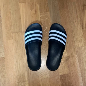 Adidas tofflor storlek 44 1/2  - Svarta Adidastofflor i storleken 44/12 men passar också mig som har storlek 44. Köpte 2 par tofflor, så dom här har varit oanvända sedan köp. Skriv ifall ni har frågor!