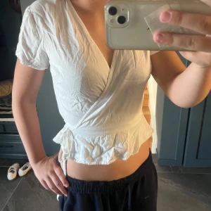 Vit omlottopp med volangkant - Säljer en söt vit omlottopp med korta ärmar och volangkant nedtill. Toppen har v-ringning och knyt i sidan för en snygg passform. Perfekt för dig som gillar en feminin och trendig stil.