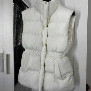 Vit dunväst från Dakfrogs, M - Säljer en fluffig vit dunväst från Dakfrogs med hög krage och markerad midja. Västen har två fickor framtill, dragkedja och tryckknappar. Perfekt för lager-på-lager och ger en clean look till din outfit.