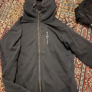 Svart hoodie från Sail Racing med dragkedja - Svart hoodie från Sail Racing med hel dragkedja och huva med snörning. Tröjan har en bröstficka med dragkedja och logga på bröstet. Muddar vid ena ärmslut och nederkant. Perfekt för dig som gillar en sportig och avslappnad stil.