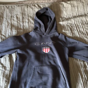 Mörkblå hoodie från GANT - Snygg mörkblå hoodie från GANT med broderad logga och sköld på bröstet. Klassisk känguruficka framtill och stor huva. Perfekt för dig som gillar stilren och sportig look. Mjuk och skön bomullskänsla. Knappt använd och inga skavanker. Jättebra skick.