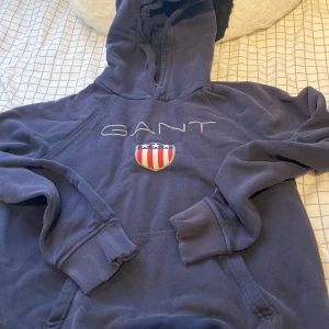 Marinblå hoodie från GANT - Säljer en marinblå hoodie från GANT med broderad logga och amerikanskt sköldmärke på bröstet. Tröjan har huva, långa ärmar och klassisk känguruficka. Perfekt för dig som gillar preppy stil och vill ha något bekvämt och snyggt.
