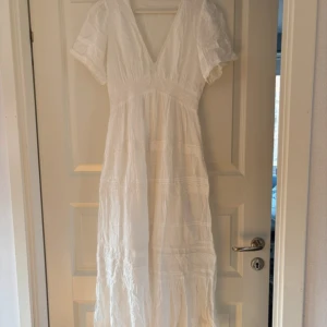 Vit långklänning med spets och prickar - Superfin vit långklänning med v-ringning och korta ärmar. Klänningen har broderade prickar och spetsdetaljer längs midjan och kjolen. Luftigt material, perfekt för varma dagar. Romantisk och bohemisk vibe, passar dig som gillar detaljer och mjuka färger.