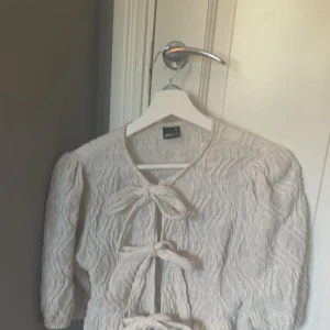 Beige blus med knyt från Gina Tricot - Superfin vit  blus från Gina Tricot med tre knyt framtill och puffiga trekvartsärmar. Blusen har en vågig struktur i tyget. Passar snyggt till både jeans och kjol. Storlek xs men passar även S 