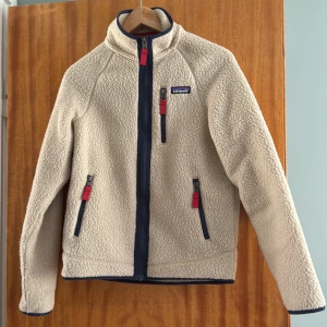 Beige fleece från Patagonia XS - Säljer en beige teddyjacka från Patagonia i storlek XS. Jackan har marinblå detaljer, röda dragkedjor och hög krage. Tillverkad i mjuk fleece som håller dig varm under kyliga dagar. Perfekt för lager-på-lager och har tre fickor med dragkedja.