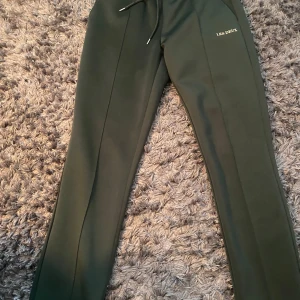 Mörkgröna trackpants från Les Deux - Snygga mörkgröna mjukisbyxor från Les Deux med diskret logga på låret. Byxorna har snörning i midjan och raka ben med en clean look. Materialet är mjukt och skönt, perfekt för chill dagar. Passformen är normal och de har en stilren design som funkar till många outfits. Dom är helt nya och har aldrig använts 