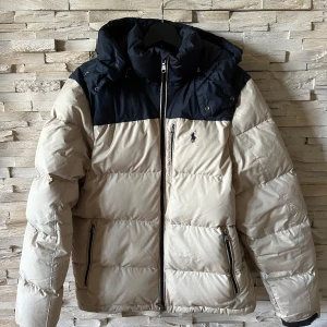 Beige och svart pufferjacka Polo Ralph Lauren - Snygg pufferjacka från Polo Ralph Lauren i beige med svart axelparti och huva. Jackan har dragkedja framtill, två fickor med dragkedja och det klassiska Polo-logot på bröstet. Perfekt för kalla dagar med sin vadderade design och sköna passform.