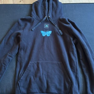 Svart hoodie med blå fjäril The Cool Elephant - Svart hoodie från The Cool Elephant med stor blå fjäril på ryggen och mindre fjäril på bröstet. Klassisk känguruficka, huva med snörning och mjukt material. Perfekt för dig som gillar unika tryck och vill sticka ut lite extra.