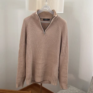 Beige stickad tröja Dressmann half zip - Beige stickad tröja från Dressmann i storlek M. Tröjan har en hög krage med half zip-dragkedja och är ribbstickad i ett grovt mönster. Långärmad och perfekt för lager-på-lager under kyliga dagar.