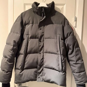 Svart pufferjacka från Canada Goose - Säljer en svart pufferjacka från Canada Goose med klassisk logga på ärmen. Jackan har hög krage, dragkedja och knappar framtill samt två stora fickor. Fylld med dun för maxad värme och perfekt för kalla dagar. Pälskrage följer också med! Passar mer som storlek m! Skriv om ni har några funderingar eller för fler bilder! ENDAST UPPHÄMTNING I MALMÖ!!