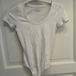 Vit ribbad bodysuit från Hollister - Supersnygg vit ribbad bodysuit från Hollister med korta ärmar och rundad urringning❤️Tight passform som sitter perfekt och knäppning nedtill. Perfekt att styla med jeans eller kjol🥰🥰