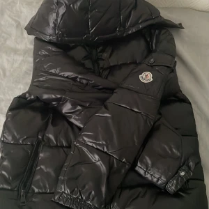 Svart Moncler dunjacka med huva - Säljer en svart Moncler dunjacka med glansig finish och huva. Jackan har dragkedja framtill, två fickor med dragkedja och klassisk Moncler-logga på ärmen. Perfekt för kalla dagar och riktigt snygg streetstil.
