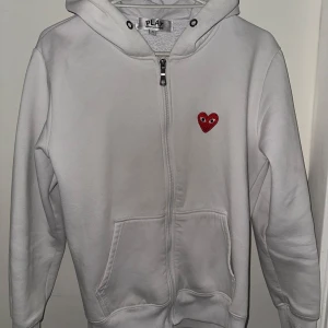 Vit hoodie Comme des Garçons Play - Vit hoodie från Comme des Garçons Play med dragkedja och huva. Klassisk röd hjärtlogga med ögon på bröstet. Mjuk bomullskänsla, känguruficka framtill och långärmad modell. Perfekt för en clean och stilren look.