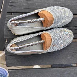 Silvriga ballerinaskor med paljetter från Duffy - Glittriga ballerinaskor från Duffy i silver med massor av paljetter på ovandelen. Skorna har rund tå och platt sula, perfekt för dig som vill sticka ut lite extra. Materialet är syntet och de har en riktigt snygg metallic-finish. (Endast skorna säljes) Passar smala fötter! 🥰