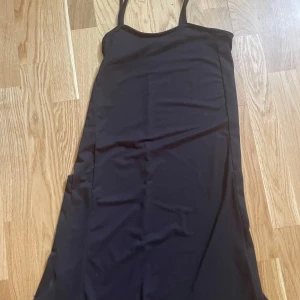 Svart långklänning med smala axelband - En stilren svart långklänning med smala axelband och rak passform. Klänningen har minimalistisk design och är perfekt för dig som gillar en clean och enkel look. Materialet känns mjukt och följsamt mot kroppen.