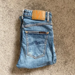 Nudie Jeans blå - Snygga ljusblå jeans från Nudie Jeans med klassisk femficksdesign och orangea sömmar. Jeansen har en rak passform och är tillverkade i mjuk bomullsdenim. Bakfickorna har Nudie Jeans signaturbrodyr och läderpatch med logga i orange. Storleken är 26/32 men passar större