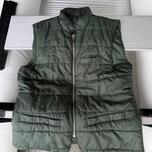 Mörkgrön dunväst från John Henric - Snygg mörkgrön dunväst från John Henric med quiltad design och hög krage. Västen har dragkedja framtill och flera praktiska fickor. Perfekt att slänga över en hoodie eller tröja för extra värme under kyliga dagar.