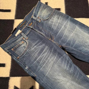 Nudie Jeans blå straight jeans 28/32 - Säljer ett par klassiska blå jeans från Nudie Jeans med snyggt slitna detaljer och kontrastsömmar. Modellen har raka ben och ikonisk brodering på bakfickorna. Tillverkade i bomull och passar perfekt till en avslappnad stil.