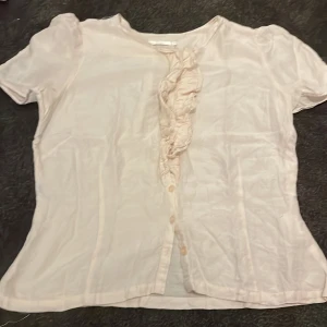 Ljusrosa blus med volang från Wera - Supersöt ljusrosa blus från Wera med korta puffärmar och knappar framtill. Blusen har en snygg volangdetalj längs knäppningen som ger en romantisk vibe. Perfekt att styla med jeans eller kjol för en fräsch look. Den är även sydd på insidan för att bli lite tajtare men är inget problem att ta bort.