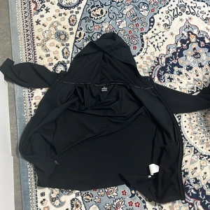 Svart vindjacka från Under Armour - Svart vindjacka från Under Armour med huva och dragkedja framtill. Jackan har en stilren look med logga på bröstet och är tillverkad i ett lätt syntetmaterial som passar perfekt för träning eller när du vill ha en sportig vibe.