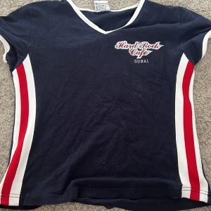Hard Rock Café Dubai t-shirt marinblå - Marinblå t-shirt från Hard Rock Café Dubai med vita och röda ränder längs sidorna, v-ringad hals och tryck både fram och bak. T-shirten är i bomull med lite stretch och har korta ärmar. Perfekt för dig som gillar klassisk rockstil.
