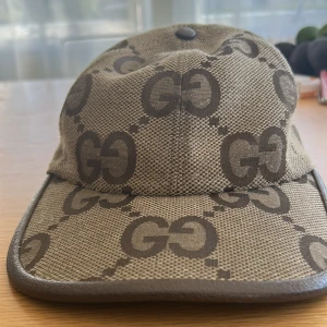 Gucci beige keps med GG-mönster - Snygg keps från Gucci i beige och brunt med det klassiska GG-monogrammet över hela kepsen. Justerbar rem baktill och detaljer i brunt läder runt skärmen. Tillverkad i bomullscanvas med exklusiv känsla och tydlig Gucci-label på insidan.