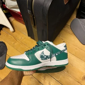 Nike Dunk Low grön och vit paisley - Nike Dunk Low sneakers i grön och vit färg med paisley-mönster på swoosh och häl. Har använt typ 2 gånger. Köpt för två år sedan.