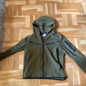 Olivgrön Nike vindjacka med huva M - Nike vindjacka i olivgrönt med hel dragkedja, huva och diskret logga på bröstet. Jackan har en snygg detalj med dragkedja på ärmen och svarta kantdetaljer. Perfekt för dig som gillar sportig och clean stil.