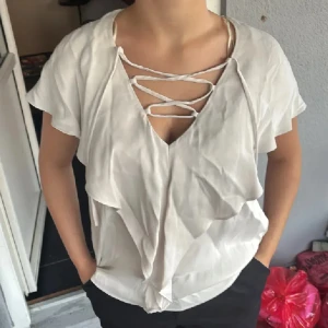 Ljusgrå blus med snörning och volanger - Supersöt ljusgrå blus med korta ärmar, djup v-ringning och snygg snörning framtill. Blusen har volangdetaljer längs framsidan som ger en flowy känsla. Perfekt för dig som gillar en feminin och trendig stil. Nyskick men behöver strykas😁 Strl 34 på den men passar xs/s