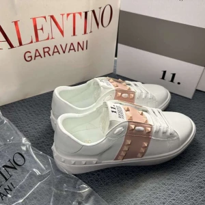 Valentino Rockstud vita sneakers - Snygga vita Valentino Rockstud sneakers med ljusrosa band och ikoniska nitar över vristen. Skorna har rund tå, platt sula och är tillverkade i skinn. Perfekta för dig som vill sticka ut med exklusiva detaljer och modern design.