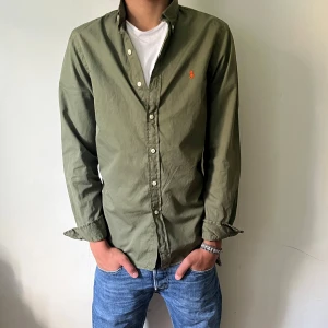 Olivgrön skjorta från Ralph Lauren - Snygg olivgrön skjorta från Ralph Lauren i slim fit med klassisk krage och knappar framtill. Materialet är lätt bomullstwill och har en orange broderad logga på bröstet. Perfekt för dig som gillar stilrena och tidlösa plagg.