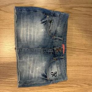 Blå jeanskjol  - Säljer en kort blå jeanskjol med slitna detaljer och rå kant nedtill. Kjolen har klassiska fickor fram och bak, dubbla knappar i midjan och coola broderade detaljer. Väldigt älskad kjol som jag köpt second hand för flera år sedan.