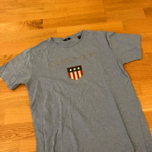 Blå GANT t-shirt med tryck - Snygg blå t-shirt från GANT med klassiskt tryck på bröstet och sköld med stjärnor och ränder. Rund hals och korta ärmar. Mjuk och skön bomull som passar perfekt till jeans eller shorts. Stilren design med GANT-logga framtill.