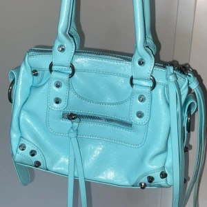 Turkos axelväska med nitar och dragkedja - Cool turkos axelväska i syntet med silvriga metalldetaljer, nitar och dragkedja framtill. Väskan har dubbla handtag, dekorativa ringar och långa remmar. Oanvänd har bara stått i garderoben. 