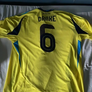 Sverige herrlandslagströja Adidas XL - Gul Sverige fotbollströja från Adidas i storlek XL, slim fit. Tröjan har blå detaljer, nummer 6 och namnet Drake på ryggen samt det svenska landslagets emblem på bröstet. Klassisk design med korta ärmar och blå ränder på axlarna. Tillverkad i lätt och ventilerande material.