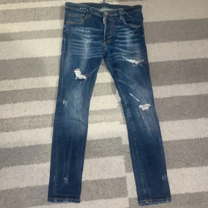 Dsquared2 blå slitna jeans - Säljer ett par blå jeans från Dsquared2 med coola slitningar och lagade detaljer. Jeansen har klassisk femficksmodell, smal passform och snyggt tvättad denim. Perfekta för dig som gillar streetstyle och vill sticka ut lite extra.