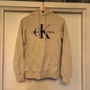 Beige hoodie från Calvin Klein Jeans - Snygg beige hoodie från Calvin Klein Jeans med stor CK-logga framtill. Tröjan har huva med snörning, känguruficka och ribbade muddar. Tillverkad i 100% bomull för en skön och avslappnad känsla. Perfekt för dig som gillar stilrena och klassiska plagg.