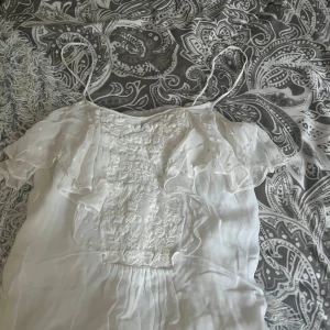 Vit topp med volang från Zara Woman - Supersöt vit blus från Zara Woman med tunna axelband, broderade blommor framtill och volangiga ärmar. Toppens material känns lätt och flowy, perfekt för varma dagar. Broderade detaljer och transparent volang ger en romantisk vibe.