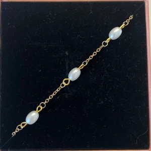 Guldarmband med vita pärlor - Snyggt armband med tunn guldfärgad kedja och tre ovala vita pärlor. Kedjan har små länkar som ger ett elegant intryck och pärlorna är jämnt placerade längs armbandet. Perfekt för dig som gillar stilrena smycken med en touch av lyx. I rostfritt stål och handgjort