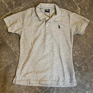 Grå pikétröja från Polo Ralph Lauren - Snygg grå pikétröja från Polo Ralph Lauren med klassisk krage, korta ärmar och broderad logga på bröstet. Tillverkad i mjuk bomull.
