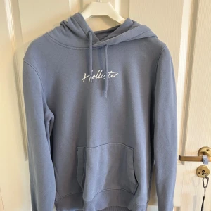 Blå hoodie från Hollister - Snygg blå hoodie från Hollister Must-Have Collection med vit broderad logga på bröstet och huvan. Tröjan har känguruficka, snörning i huvan och är i mjuk bomullsblandning. Perfekt för chill dagar och streetstyle. Är ett set med mjukisbyxorna.