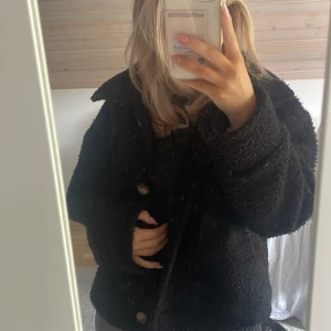 Svart teddyjacka från Chelsea Girls - Mysig svart teddyjacka från Chelsea med stora knappar framtill och bred krage. Jackan har en boxig passform och är perfekt för kyligare dagar. Materialet är fluffigt och ger en avslappnad vibe. Passar dig som gillar trendiga och bekväma ytterplagg.