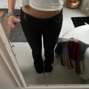 Svarta bootcut jeans med låg midja - Säljer ett par svarta jeans med låg midja och bootcut-snitt. Jeansen har klassisk femficksdesign, snygga detaljer med dubbla knappar i midjan och är tillverkade i ett stretchigt bomullsmaterial. Perfekta för dig som gillar en tidlös och clean look.