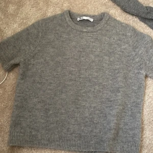 Grå stickad tröja från Zara - Mysig grå stickad tröja från Zara med korta ärmar och rund halsringning. Tröjan har en enkel och clean look med ribbade kanter vid hals, ärmslut och nederkant. Perfekt för lager-på-lager och chill vibes.