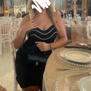 Svart kort klänning med strassdetaljer - Supersnygg svart  klänning med tunna axelband och vågformade strassdetaljer längs bysten. Klänningen har figurnära passform och dragkedja i ryggen. Perfekt för dig som vill sticka ut med en elegant och trendig look. Används en gång